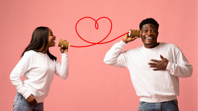 Two joyful African American lovers communicate through a tin can phone, sharing words of love and fun on Valentine's Day, with a bright pink studio background enhancing the festive mood.