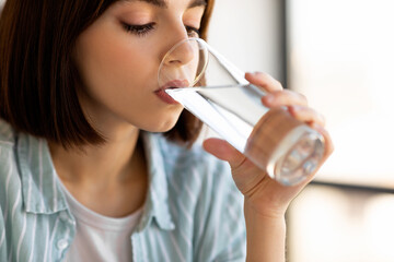 Fototapeta premium A young woman with short hair enjoys a refreshing drink of water from a glass. She sits peacefully indoors, illuminated by natural light.