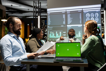 Diverse project team meets after hours next to mockup display, sharing data visualization reports to discuss business intel. Preparing forecasts for upcoming presentation to investors group.