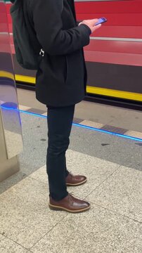 platform standing man checking phone near red train, casual jacket and jeans, brown shoes on tiled platform, scrolling screen, waiting posture. Vertical, taken on the mobile device.