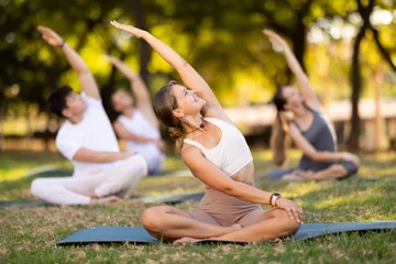 Fotobehang Lotusbloem Fit young girl enjoying group yoga session in peaceful environment of verdant summer park, performing stretches in lotus position while sitting on exercise mat  © JackF