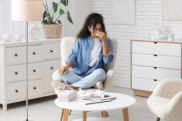 Young African-American woman sneezing in handkerchief and sitting on armchair at home. Allergy...