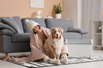 Young woman in winter clothes with blanket and Australian Shepherd dog sitting on floor near heater at home