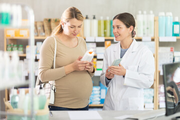 Obraz premium Interested pregnant woman reading label on stretch mark cream bottle, focusing on natural ingredients, while friendly female pharmacist providing guidance on safe skincare in background at drugstore