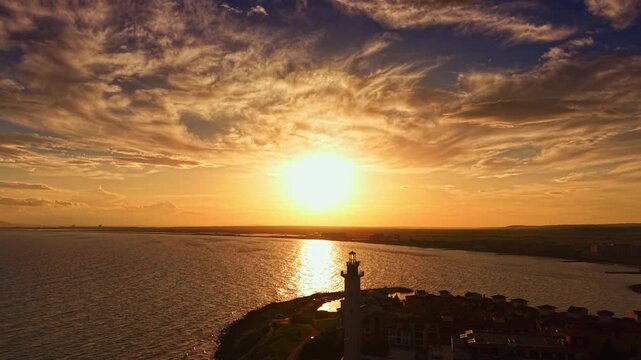 A stunning sunset envelops the coastline in vibrant hues, as the sun sinks low, creating shimmering reflections on the calm water. The lighthouse stands proud against the colorful sky.