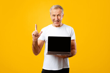 Man stands against a yellow background and smiles while holding a laptop in one hand and raising his index finger. He appears to be sharing helpful information or ideas.