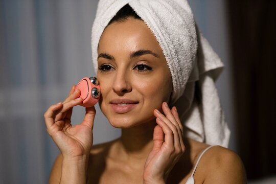Close up of Caucasian young attractive woman with towel on head using microcurrent facial toning device at home. Beauty device. Beautiful happy female using skincare device