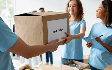 A male volunteer gives a box labeled donations to a woman who is a social worker taking notes. Colleagues are engaged in various tasks at the charity center, creating a busy atmosphere.