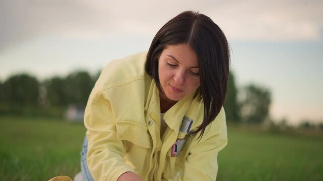 woman in yellow jacket bending over in grassy field at sunset, inspecting and gathering pumpkins with focused expression and braided hair, casual jeans and hands covered in soil, calm rural atmosphere
