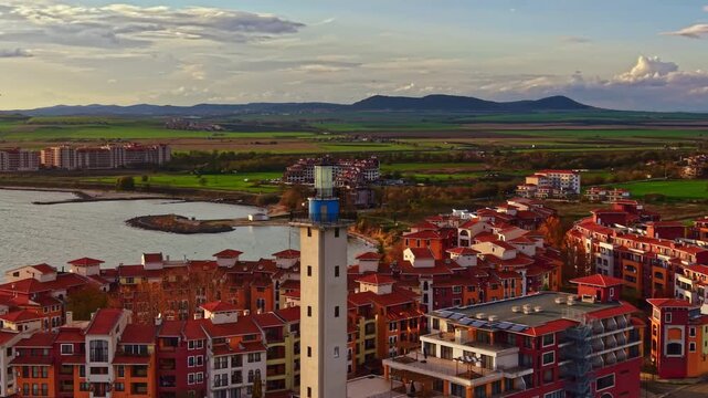 Golden rays reflect off calm waters as a lighthouse stands tall amidst colorful buildings, offering a picturesque view of the tranquil coastal town at dusk.