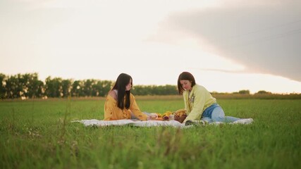 sunset field picnic two women chatting, college friends sharing fruit on blanket with gentle laughter, relaxed posture, wide grassy meadow, soft golden light over countryside horizon, casual denim