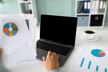 A female financial manager is focused on her laptop with a blank screen while organizing paperwork. She holds documents with diagrams, sitting in a bright office space with a cup of coffee nearby.