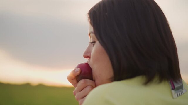 white woman profile at sunset, taking slow bite of apple during golden hour soft backlit silhouette, subtle flare over meadow, contemplative expression, student pausing study, writer reflecting
