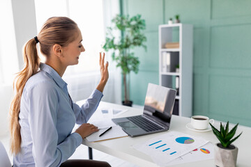 A female entrepreneur sits at a desk in her office, using a laptop to connect with her business team. She waves at the webcam, showing interaction during remote work.