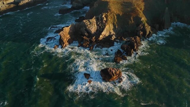 Aerial drone gliding along curved path birds-eye view above rocky coastline with crashing waves and greenish ocean water on the northern coast of Spain