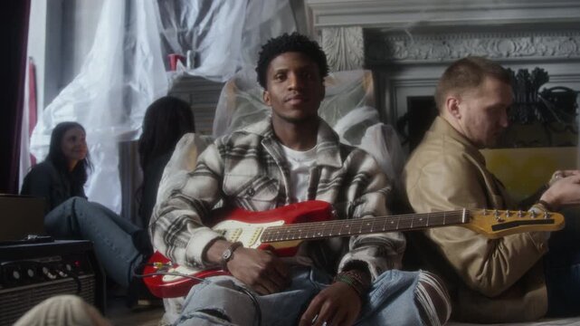 Front view shot of African American man sitting on floor in abandoned house and holding guitar while looking at camera with group of people in background, copy space