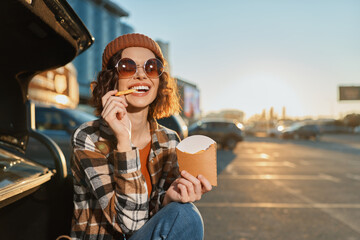 Woman eating fries by car in parking at sunset, smile with sunglasses and beanie in golden hour glow, candid lifestyle portrait conveying authenticity, mindful living and emotional storytelling.