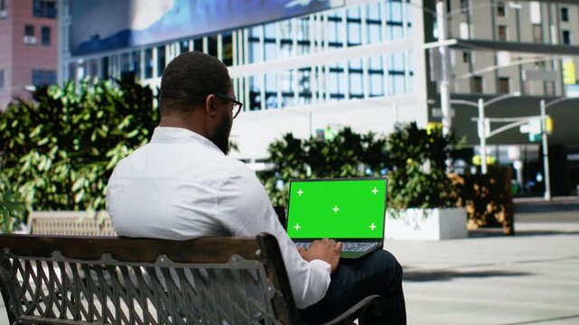 Mockup on screen with hardworking adult man uses laptop on a bench, navigating web content with steady motion and casual confidence, surrounded by architecture and vibrant urban life. Camera B.