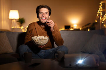 A young man smiles as he watches a movie using a projector in his cozy living room. He enjoys a bowl of popcorn while relaxing on the sofa during a weekend evening.
