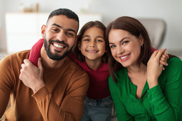 A cheerful Middle Eastern family of three sits on a sofa, smiling and hugging each other. The young girl joyfully embraces her parents, revealing a loving home atmosphere filled with happiness.