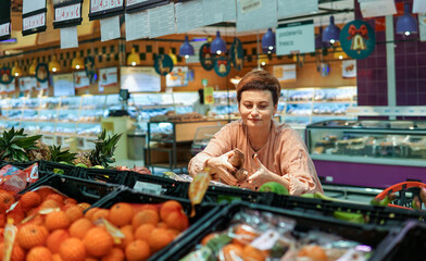 Obraz premium Middle-aged woman in pink suit choosing fruits and vegetables in a large shopping mall supermarket