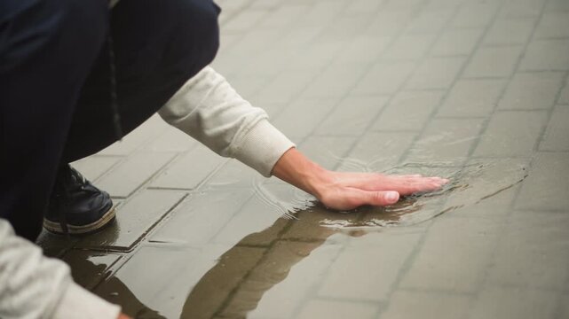 asian man crouching on wet pavement scooping rainwater from puddle, sequence shows hand skimming tiled surface, splash and ripple details, wet hair and glasses dripping, candid street portrait