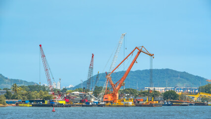 Large industrial cranes, including an orange gantry crane, are situated on barges and waterfront, set against a blue sky and distant green hills