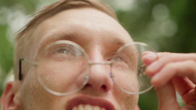 young white man clear glasses upward with hopeful smile, droplets fading, sunlight filtering through leaves, green bokeh background, closeup cinematic perspective, renewed clarity and relief. Frame