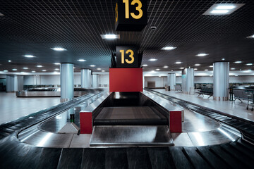 Low-key interior of empty airport baggage claim hall with luggage carousel, dramatic lighting, symmetry and perspective, conveyor belt and numbered sign, modern transport terminal atmosphere
