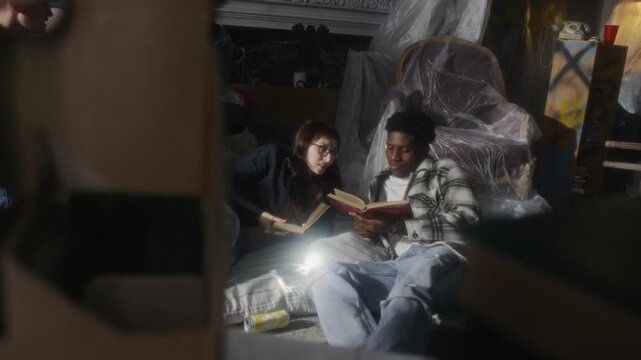 Authentic sneak shot of African American man talking to young woman while sitting on mattress and hanging out with group of friends in abandoned house with furniture in foreground, copy space