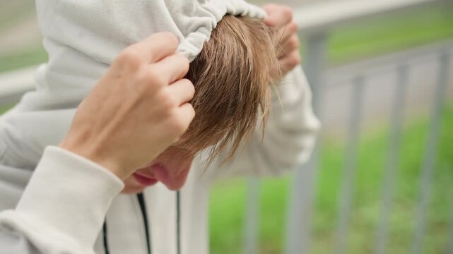 closeup white male pulling hood over face, park railing, rainy cloudy day, hands adjusting fleece jacket, wet hair, anxious student seeking shelter, intimate profile showing vulnerability and tension