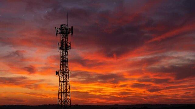 A lone cell tower stands in silhouette against a dramatic sunset sky painted with vibrant orange and red hues and wispy clouds