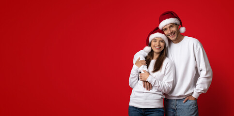 Fototapeta premium A cheerful couple stands together, wearing matching white sweaters and festive Santa hats. They smile warmly while posing against a vibrant red background, embodying the holiday spirit.