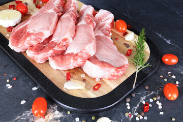 Fresh raw pork steaks on a wooden board with garlic, rosemary, and tomatoes ready for cooking on a dark table.