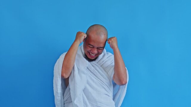 Excited Muslim pilgrim in Ihram laughing and celebrating with raised fists on blue background
