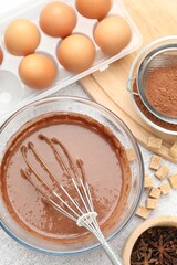 Chocolate dough with whisk in bowl, eggs and spices on grey textured table, flat lay
