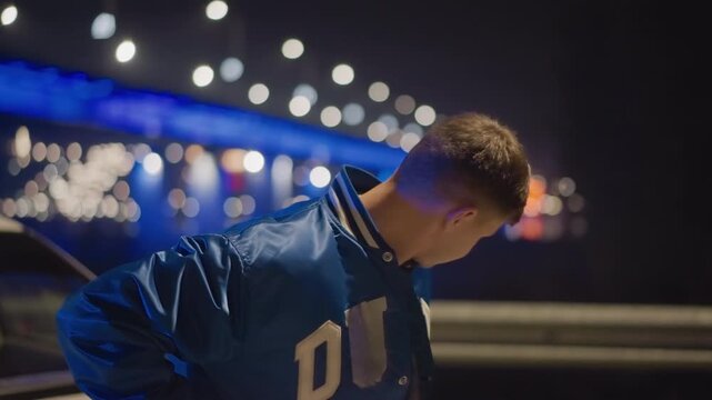 Night bridge portrait white driver near car, blue illuminated bridge lights and bokeh, contemplative young man in varsity jacket, waterfront reflections and city glow, cinematic urban atmosphere,