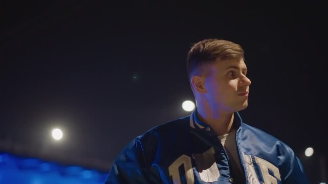 young white lookout scanning highway under blue bridge lights, tense posture and shifting gaze across lanes, checking surroundings near parked car, neon reflections on varsity jacket, cinematic patrol