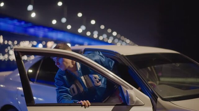 young man in car under bridge illuminated by blue lights, white sedan parked at roadside, varsity jacketed driver looks out window then steps out, surveys highway with contemplative posture, cinematic