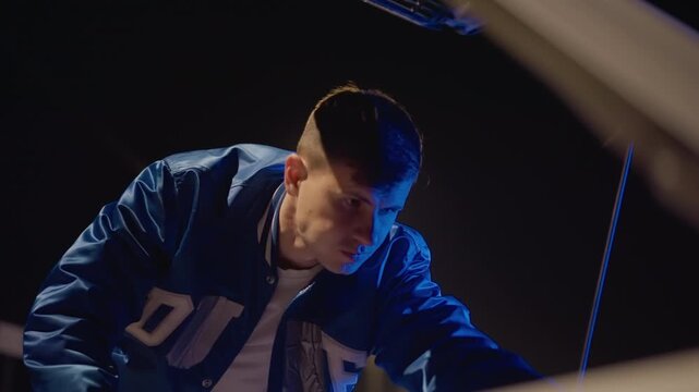 night driver inspecting car hood under blue streetlight, young man leaning into engine compartment, letterman jacket catching cool highlights, closeup cinematic angles, focused expression, urban