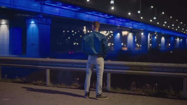 man at bluelit bridge at night, standing by guardrail overlooking river, illuminated underside of bridge with deep blue LEDs, city lights and bokeh in background, denim jacket and casual jeans,