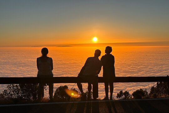 Three people watching sunset above clouds, together and alone