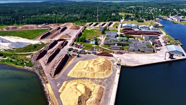 Aerial view of a coastal industrial yard filled with large piles of lumber and sawdust, machinery, storage rows, and a loading dock beside calm blue water.