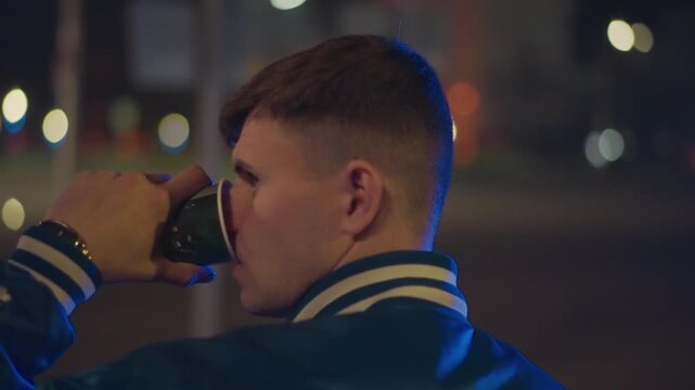 Youthful man reflecting, White male studies in evening glow, Male in jacket pauses alone on deserted night street, Young man dressed in varsity jacket stands in silent parking lot under rim light