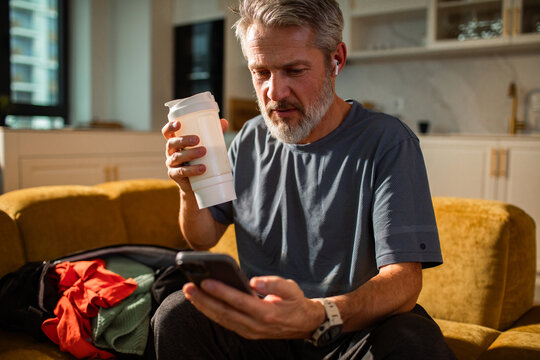 Mature man focused on smartphone with shaker on home couch