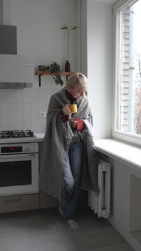 Middle aged woman stands beside window in cold apartment covered in plaid and trying to keep warm in unheated house. Female with mug of hot tea checking touching radiator, central heating problems.
