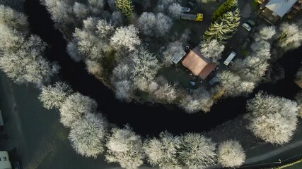Top down morning view of a small Estonian neighbourhood with a dark river, frosty trees and rooftops as sunrise light melts hoarfrost during early winter.