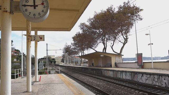 Slow downward shot of Caxias train station platform on the Portuguese coast with vintage clock, old railway tracks, and traditional stone pavement with no people present.