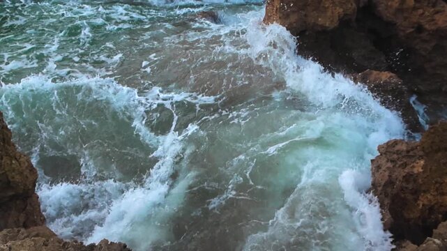 Powerful ocean waves crash against rugged rocky coastline in Portugal as white foam and turquoise water splash between stones during high tide on a cloudy day.