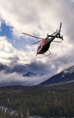 Fototapeta na wymiar Red Helicopter In Flight Over Snowy BC Mountains With Blue Sky And Dramatic Clouds